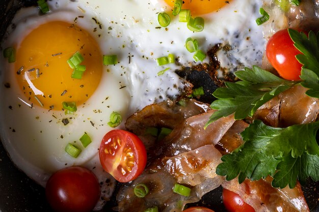 Petit-déjeuner avec des œufs au plat dans une poêle avec du bacon et des légumes sur fond sombre.