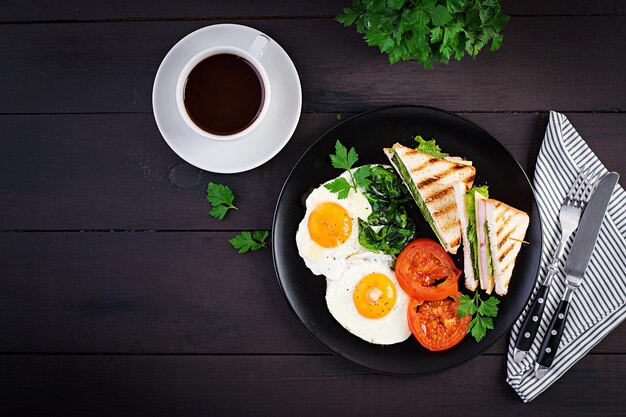 Petit déjeuner avec des œufs frits, des épinards, des tomates et un sandwich au club sur l'assiette.