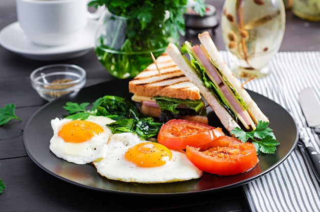 Le petit déjeuner, des œufs frits, des tomates, des épinards et un sandwich au club sur une assiette.