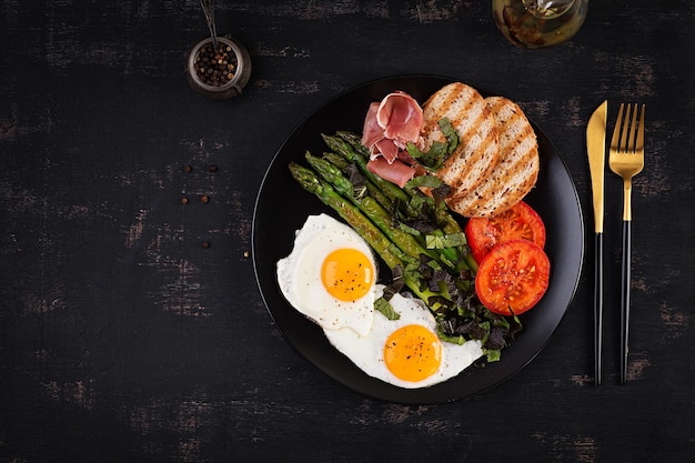 Petit-déjeuner pain aux œufs frits toasts asperges vertes tomates et jamon sur plaque noire vue de dessus ci-dessus