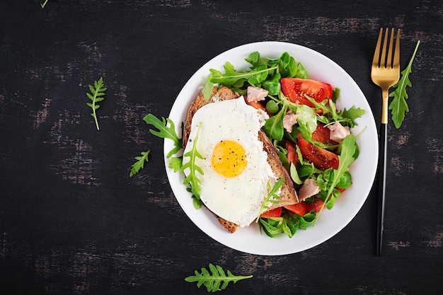 Petit-déjeuner - pain grillé avec œufs au plat et salade de tomates avec légumes verts et foie de morue sur fond sombre. Petit-déjeuner sain. Vue de dessus