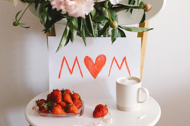 Petit-déjeuner pour la fête des mères Assiette blanche en forme de coeur avec fraises fraîches tasse de café cadeau et bouquet de pivoines avec cadeau au lit Composition nature morte Bonne fête des mères