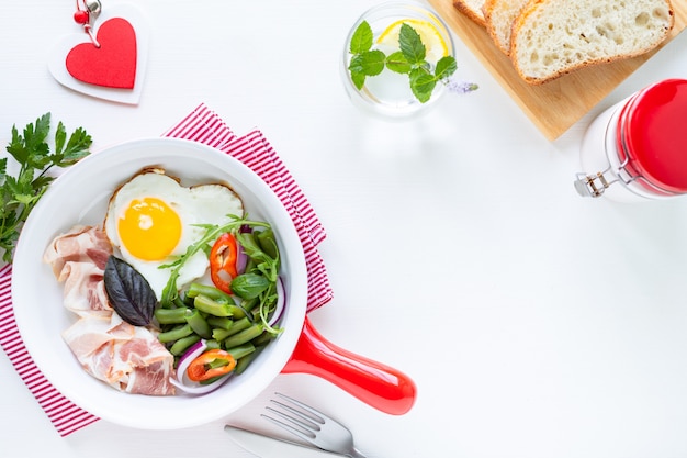 Petit-déjeuner pour votre bien-aimé pour les vacances: œuf en forme de cœur, bacon, haricots verts sur une table blanche. Mise au point sélective. Vue d'en-haut. Copiez l'espace.