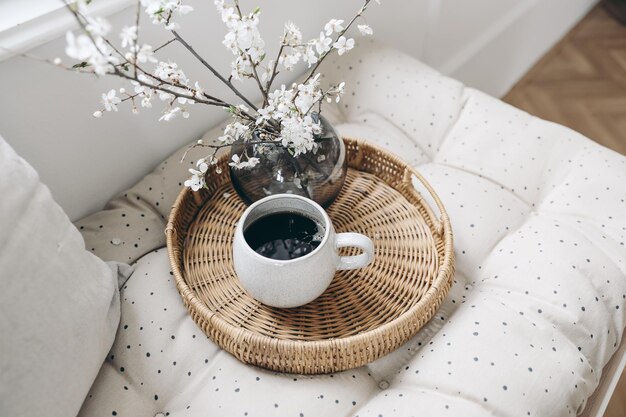 Petit-déjeuner de printemps composition de nature morte Tasse de café et plateau en osier sur matelas français Photo de style féminin Scène florale avec des fleurs de cerisier blanches floues Banc près de la fenêtre Mise au point sélective