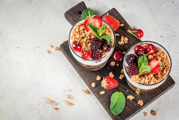 Petit-déjeuner sain. Baies et fruits d'été. Yaourt grec fait maison avec granola, mûres, fraises, cerises et menthe. Sur table en pierre de béton blanc, dans des verres. vue de dessus