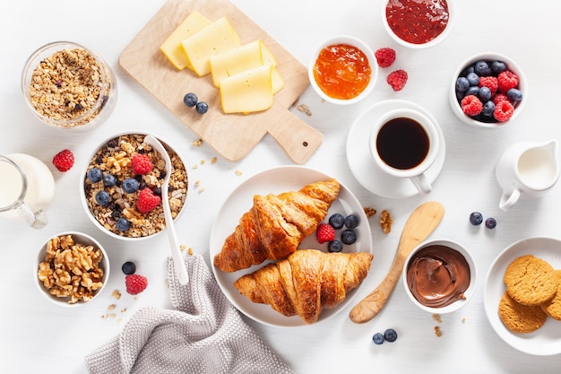 Petit-déjeuner sain avec granola, petits fruits, noix, croissant, confiture, pâte à tartiner au chocolat et café. Vue de dessus