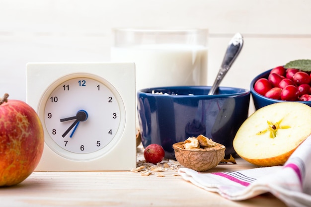 Petit-déjeuner sain - horloge, lait, flocons d'avoine, canneberges, noix, pommes sur une table en bois