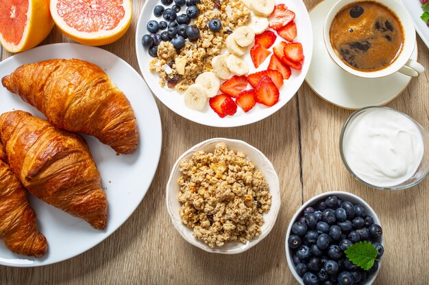 Petit-déjeuner sain servi avec assiette de yaourt muesli myrtilles fraises et banane. Table du matin granola amandes baies jus d'agrumes café croissants et herbes vertes.