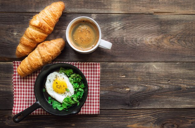 Petit-déjeuner traditionnel avec café, croissants et œuf au plat dans une poêle en fer sur bois rustique.