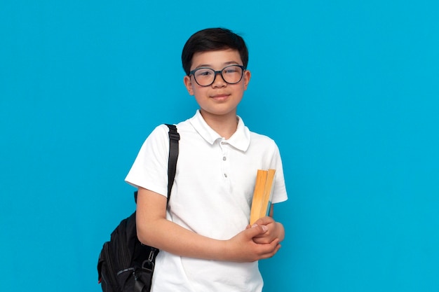 Un petit écolier asiatique en lunettes avec un sac à dos tenant des livres et souriant sur un fond bleu