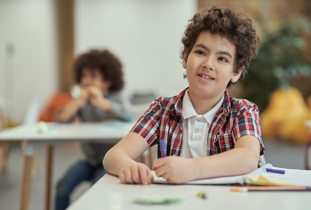 Petit écolier diligent écoutant son professeur assis au bureau dans la salle de classe