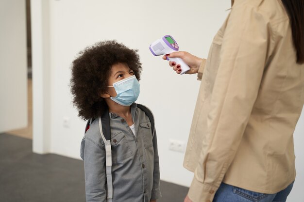 Un petit écolier espiègle portant un masque facial attend pendant que son professeur mesure la température