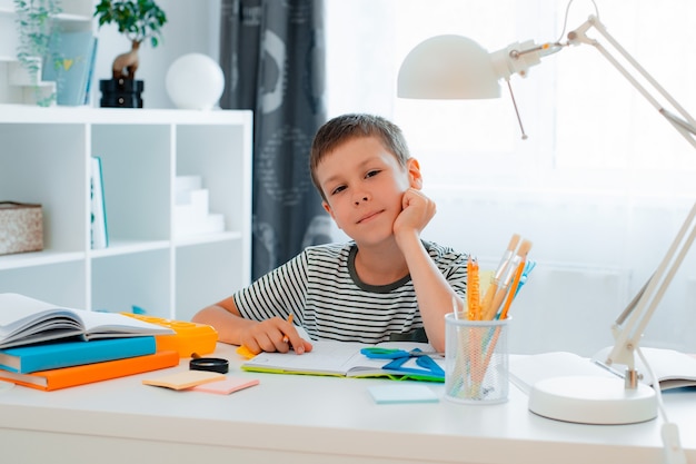 Un petit écolier est assis à la maison à la table et résout ses devoirs. Retour à l'école, préparation, enseignement à domicile.