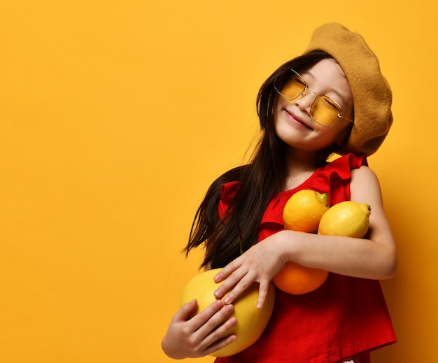 Petit enfant asiatique en lunettes de soleil, béret marron, chemisier rouge. Souriant les yeux fermés, tenant du pomelo, de l'orange et des citrons dans ses mains, posant en studio. Enfance, fruits, émotions. Gros plan, espace de copie