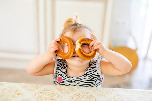 Petit enfant en bas âge en train de déjeuner dans la cuisine chaude et ensoleillée. Fille blonde avec queue de cheval drôle jouant avec deux délicieux bagels.