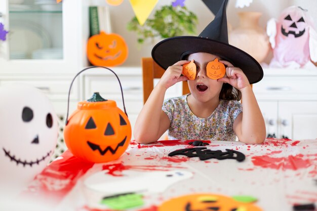 Petit enfant caucasien au chapeau de sorcière s'amusant tout en faisant des citrouilles d'Halloween à la main. Elle est dans le salon.