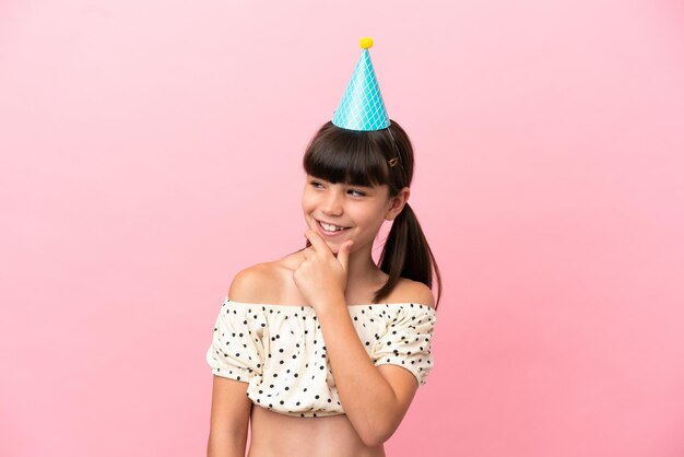 Petit enfant caucasien avec chapeau d'anniversaire isolé sur fond rose regardant sur le côté et souriant