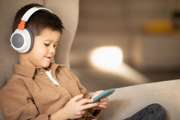 Un petit enfant caucasien souriant dans un casque sans fil décontracté regarde la vidéo sur le téléphone est assis dans un fauteuil dans le salon
