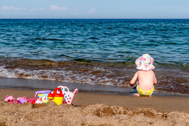 Petit enfant en chapeau et maillot de bain jouant avec des jouets sur la plage de sable