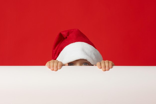 Petit enfant dans un bonnet de Noel rouge caché derrière une bannière blanche par peur