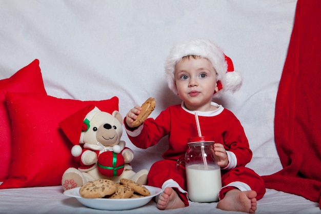 Un petit enfant dans un bonnet rouge mange des biscuits et du lait. Portrait de Noël d'un bébé dans un bonnet rouge.