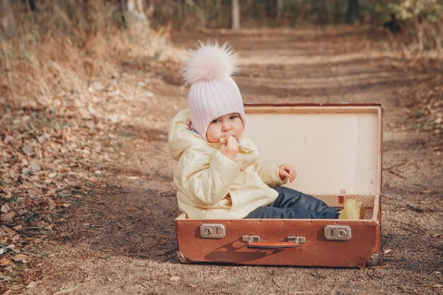 Un petit enfant dans un costume chaud est assis dans une valise sur la route. Le concept de changement de résidence, de déménagement, d'accessoires, d'air pur, de sans-abri, de maisons d'enfants