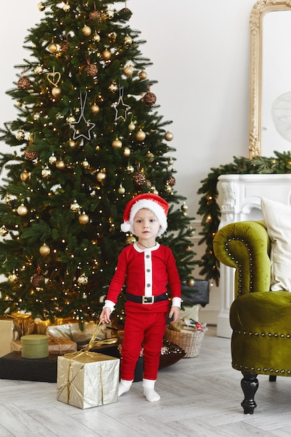 Un petit enfant dans un costume de père Noël posant avec une boîte-cadeau de Noël près de l'arbre de Noël