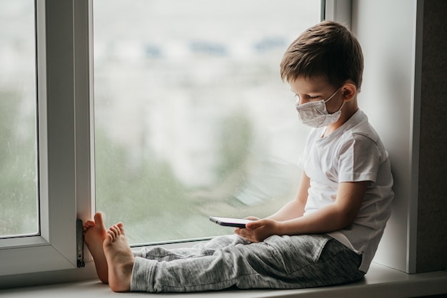 Un petit enfant dans un masque médical est placé en quarantaine à la maison sur une fenêtre avec un téléphone à la main.Prévention du coronavirus et de Covid - 19