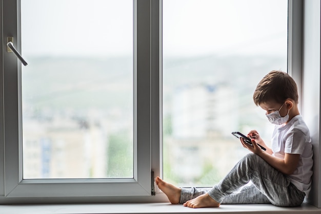 Un petit enfant dans un masque médical est placé en quarantaine à la maison sur une fenêtre avec un téléphone à la main.Prévention du coronavirus et de Covid - 19