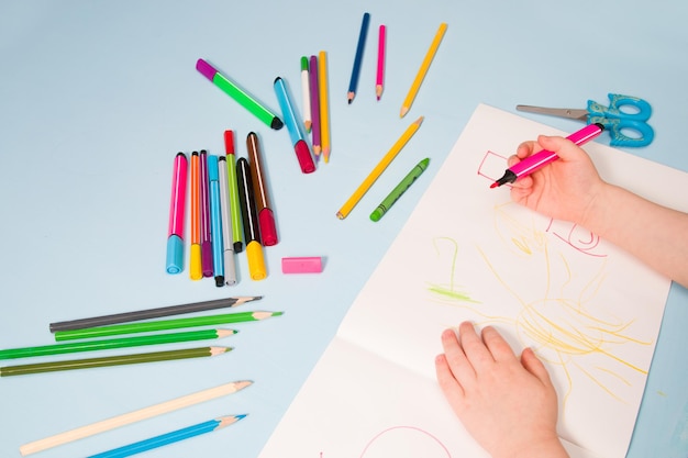 Un petit enfant dessine avec des crayons dans un album, un espace de copie, une vue de dessus, un fond bleu, des crayons, des crayons de cire, des ciseaux et les mains de l'enfant sur la table