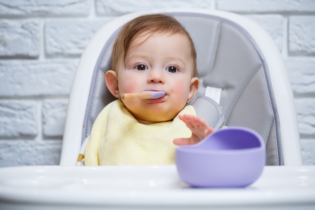 Un petit enfant est assis sur une chaise haute et mange de la nourriture dans une assiette avec une cuillère. Ustensiles en silicone pour bébé pour nourrir les bébés