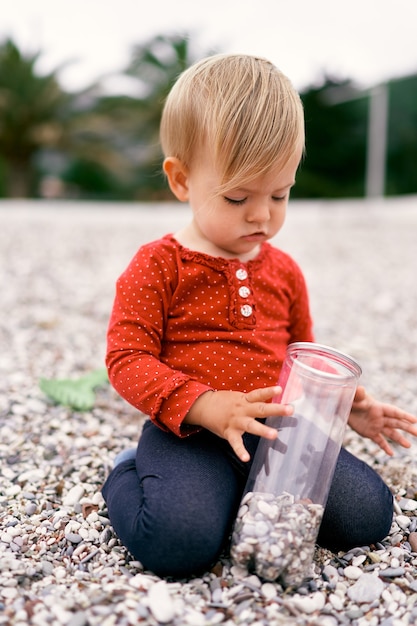 Un petit enfant est assis à genoux sur une plage de galets et tient un pot de galets dans ses mains