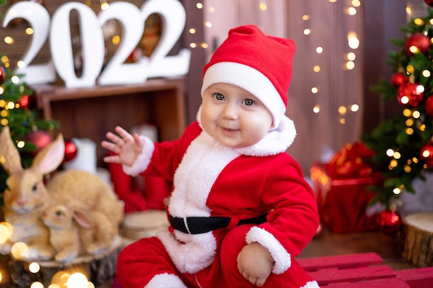 Un petit enfant est assis sur un traîneau sous les arbres de Noël. Un grand portrait. Un bébé dans un costume de Père Noël rouge avec un cadeau dans une boîte cadeau. Joyeuses Fêtes, Nouvel An. Période de Noël