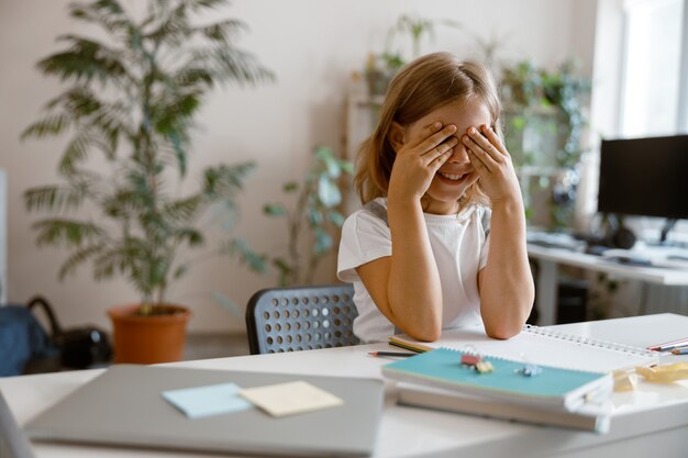 Le petit enfant ferme les yeux par les mains assis à table avec des fournitures et un ordinateur portable dans la chambre