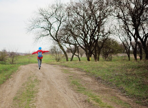 Un petit enfant un garçon dans une veste rouge et une casquette bleue saute joyeusement et joyeusement et saute sur la route dans le parc ou dans la forêt. Le concept de l'enfance, du printemps.