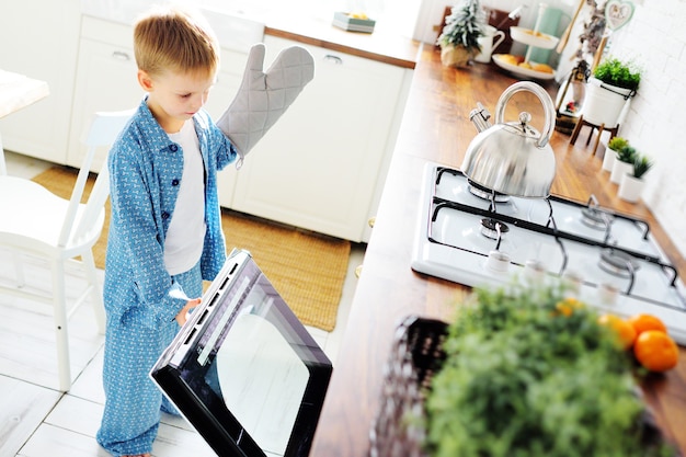 Un petit enfant un garçon en pyjama bleu avec un gant se dresse dans le contexte d'un four ouvert et sourit dans le contexte de la cuisine
