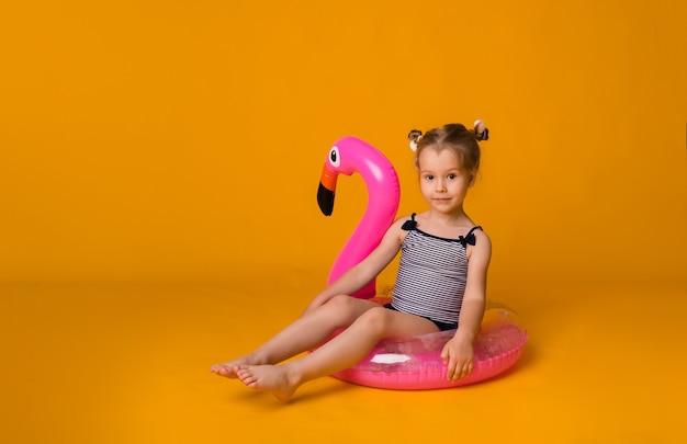 Le petit enfant mignon dans un maillot de bain rayé se repose dans un cercle gonflable rose et regarde la caméra sur un fond jaune avec l'espace pour le texte