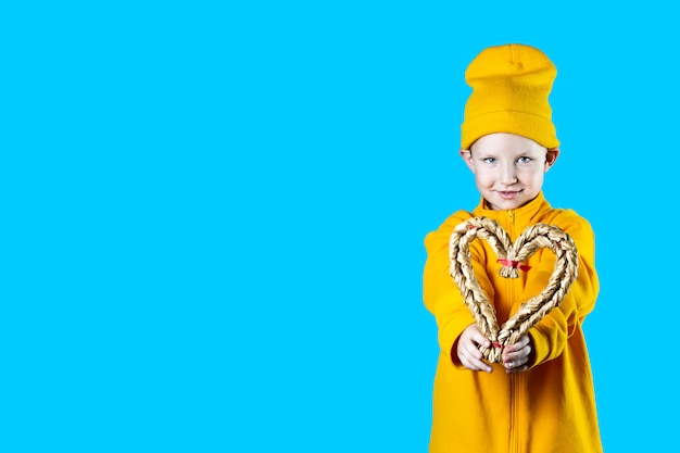 Un petit enfant mignon dans une veste jaune et un chapeau. Tenant un coeur tressé sur fond coloré.