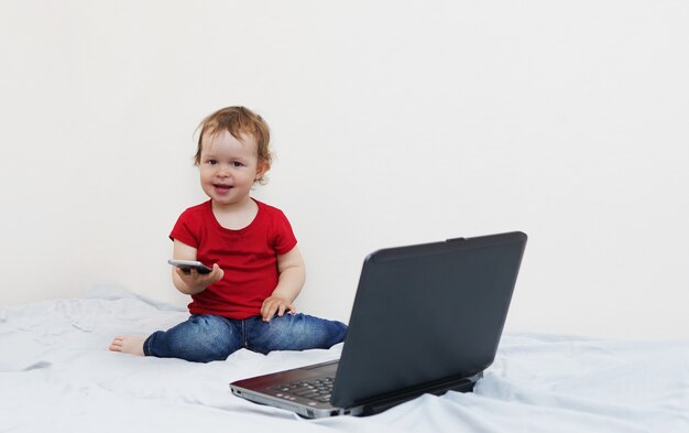 Petit enfant moderne assis sur le lit avec smartphone dans ses mains et utilisant un ordinateur portable