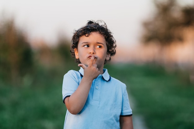 Petit enfant pensif en polo bleu touchant ses lèvres par l'index regarde de côté en pensant Mignon enfant hispanique bouclé se promène dans le parc au coucher du soleil au parc Enfance et beaux enfants Enfant drôle