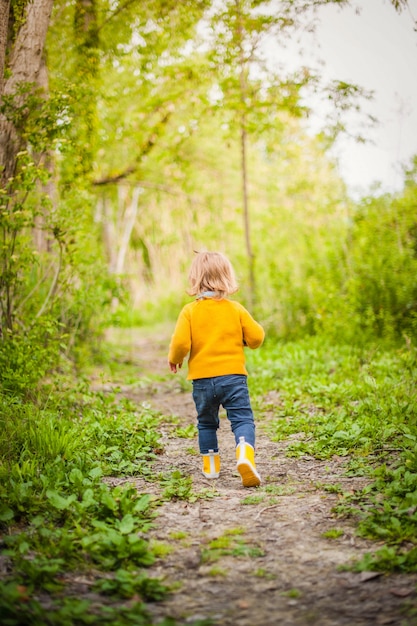 Photo petit enfant portant des bottes de pluie jaunes, marchant le long d'un chemin forestier dans l'herbe