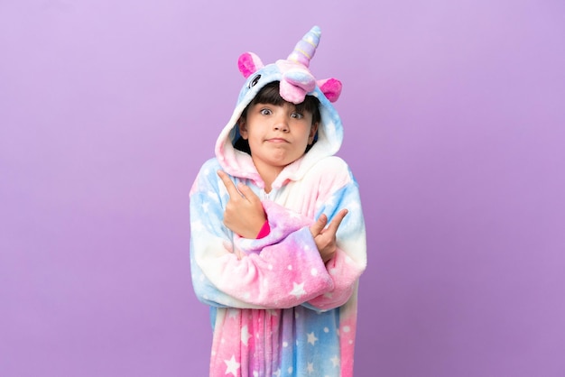 Petit enfant portant un pyjama licorne isolé sur fond violet pointant vers les latéraux ayant des doutes