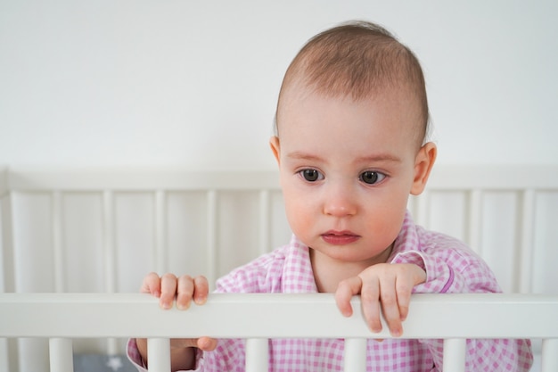 Un petit enfant en pyjama rose est debout dans le berceau. L'enfant s'est réveillé et attend que ses parents le sortent du berceau. Bébé commence à pleurer