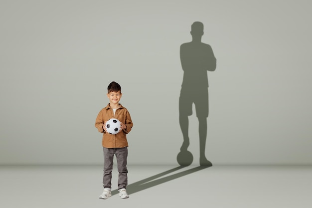 Un petit enfant de race blanche positif en tenue décontractée avec un ballon de football et l'ombre d'un jeune footballeur