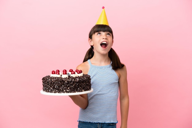 Petit enfant de race blanche tenant un gâteau d'anniversaire isolé sur fond rose en levant et avec une expression surprise