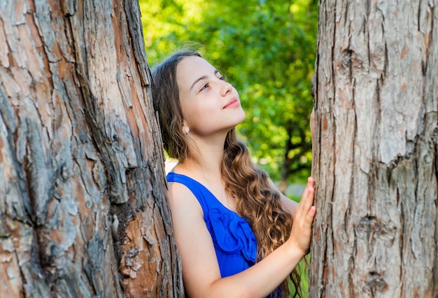 Petit enfant rêveur aux longs cheveux ondulés et au look beauté rêver au tronc d'arbre dans le parc d'été, futur.