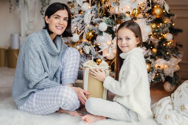 Un petit enfant et sa mère sont assis sur un tapis blanc chaud près d'un arbre du Nouvel An décoré. Ils ont des expressions heureuses et heureuses de passer des vacances ensemble. Enfance et vacances