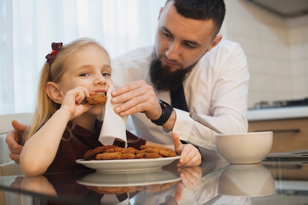 Petit enfant et son père mangeant des biscuits à la cuisine pendant que le père nettoie son visage. Père et fille le matin à manger dans la cuisine.