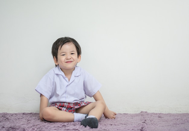 petit enfant en uniforme d&#39;étudiant avec le visage de sourire s&#39;asseoir sur un mur et un mur blanc avec fond