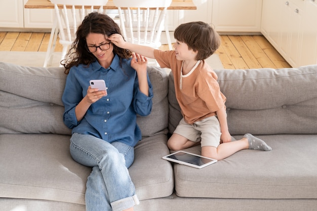 Un petit fils actif qui s'ennuie distrait une jeune femme d'affaires mère qui travaille en ligne sur un smartphone depuis la maison